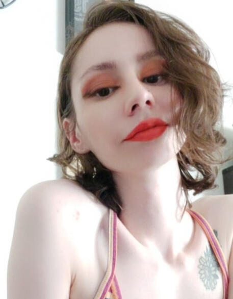 a selfie of Acey wearing a striped camisole, red lipstick, pink eyeshadow & angled black eyeliner, she has short curly brown hair and a sunflower tattoo under her left collarbone